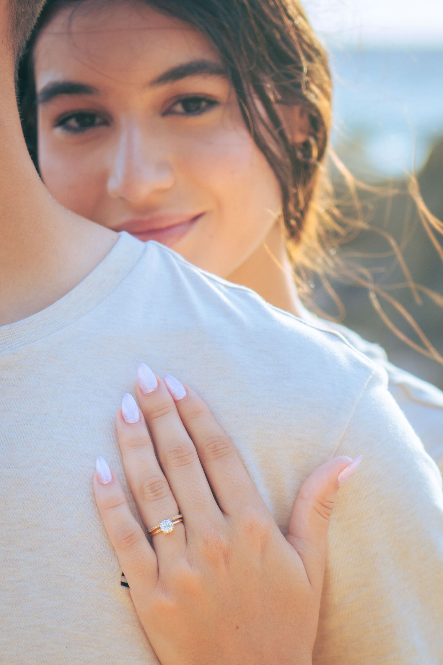 Woman smiling and resting her hand on a man's chest, showing off a delicate diamond engagement ring.