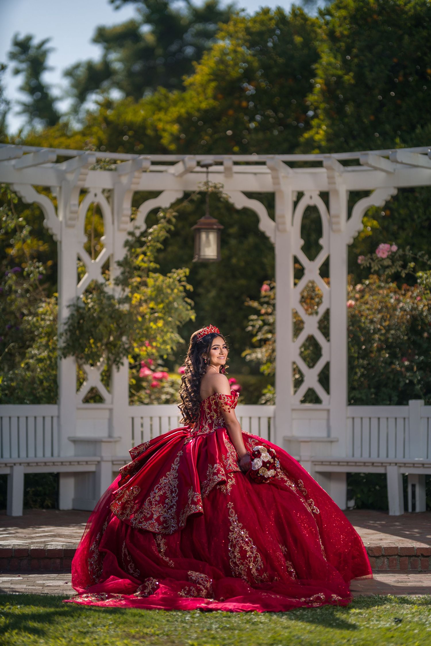 Young woman in a stunning red and gold ball gown poses in a white garden gazebo surrounded by lush greenery.