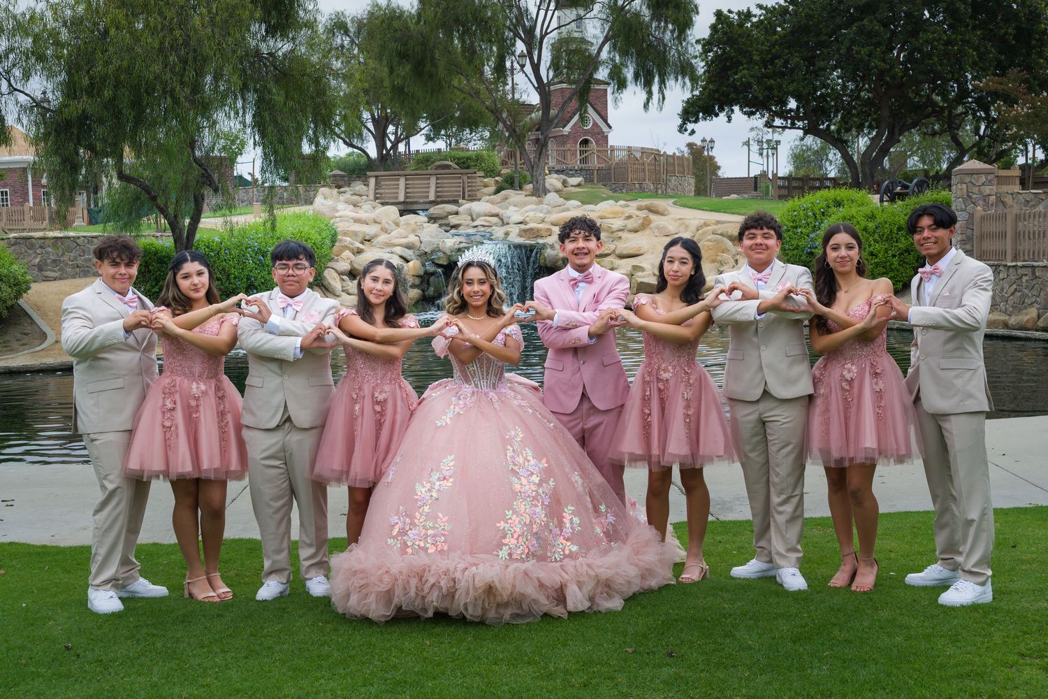 Quinceañera celebration with court in pink dresses and tan suits posing outdoors near a pond.
