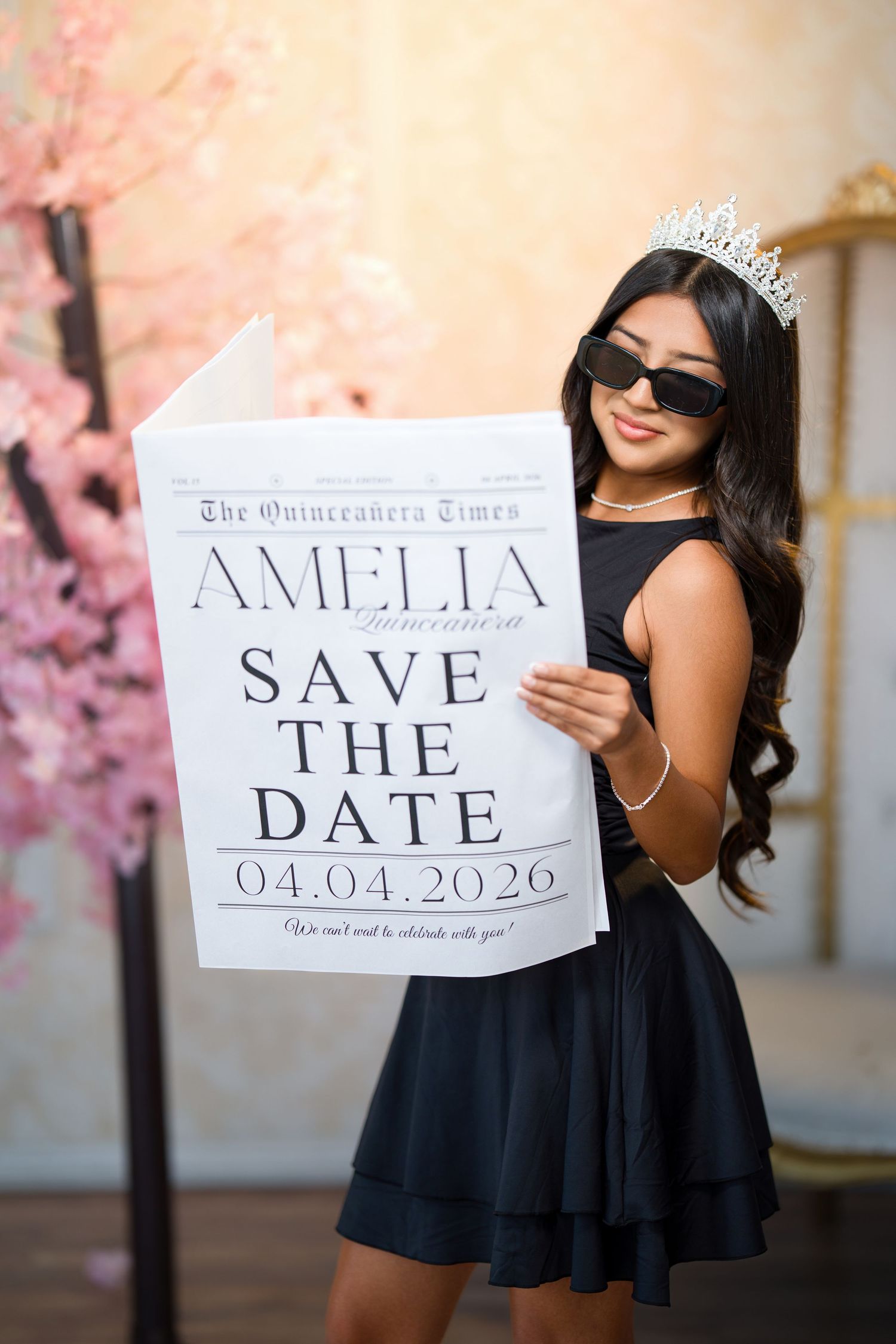 Quinceañera girl in a black dress, tiara and sunglasses holds a newspaper-style save the date for Amelia, April 4, 2026.