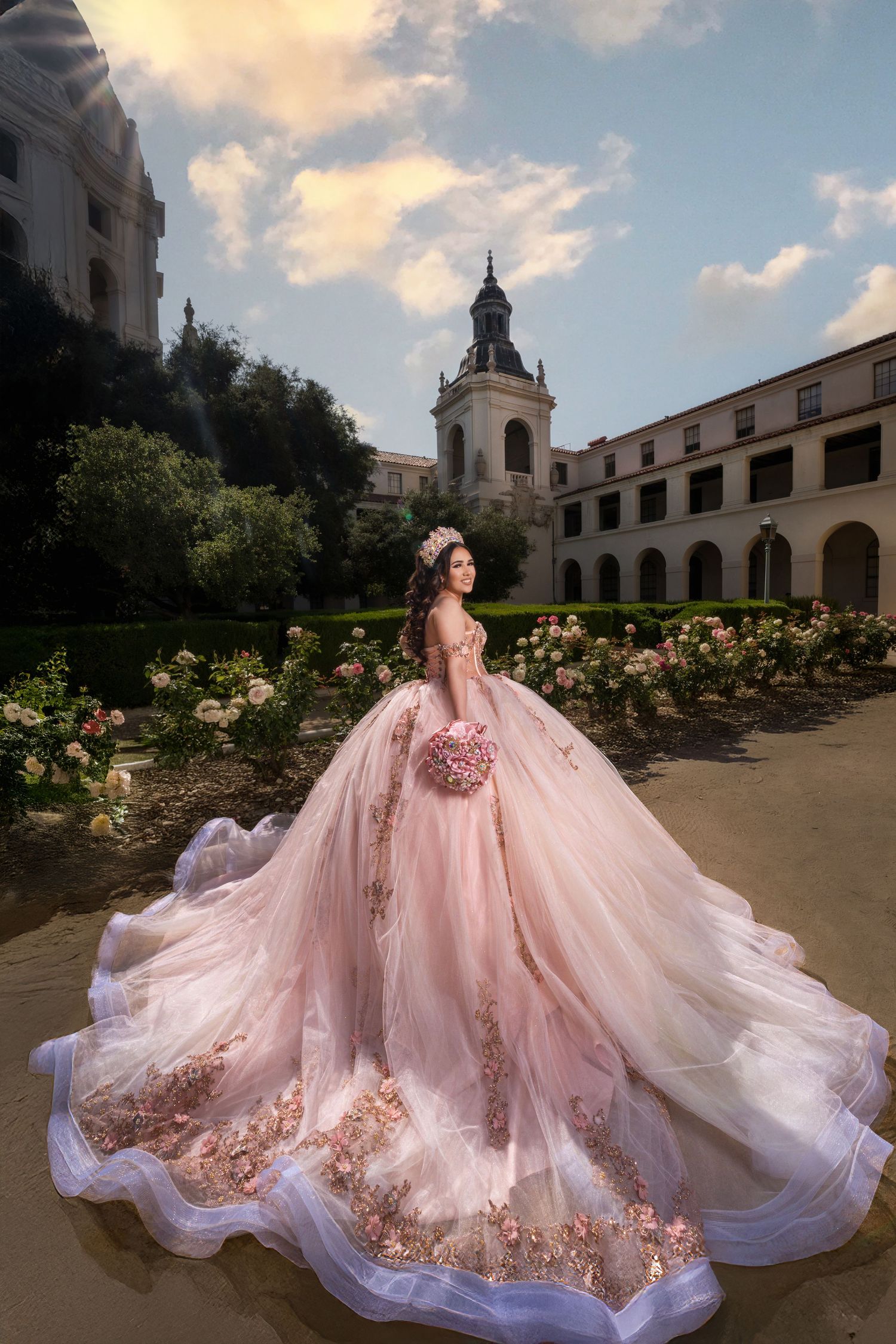 Elegant blush pink ball gown with long train and floral details photographed in courtyard with church tower in background.