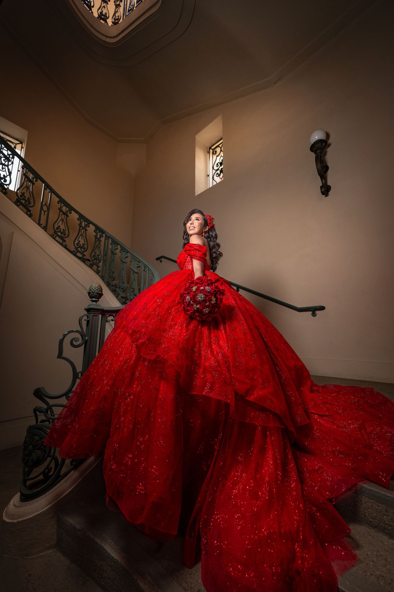 A stunning red ball gown with a voluminous tulle skirt cascades down an elegant staircase with wrought iron railings.