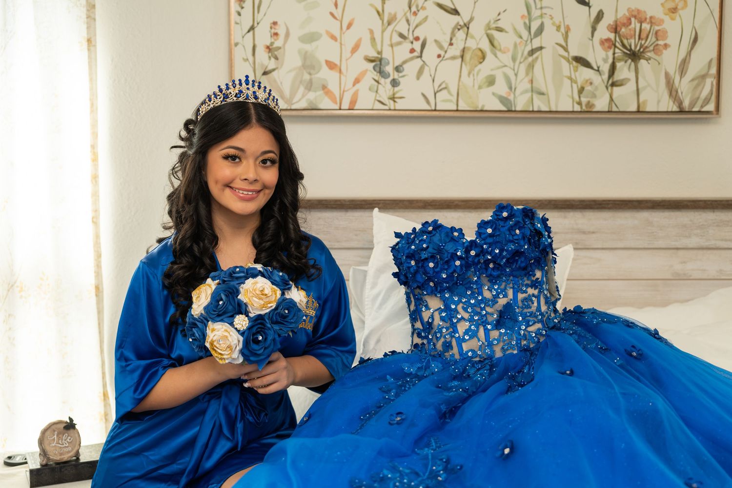 Young Latina girl in royal blue robe holding bouquet beside elegant blue quinceañera ball gown on bed with tiara.