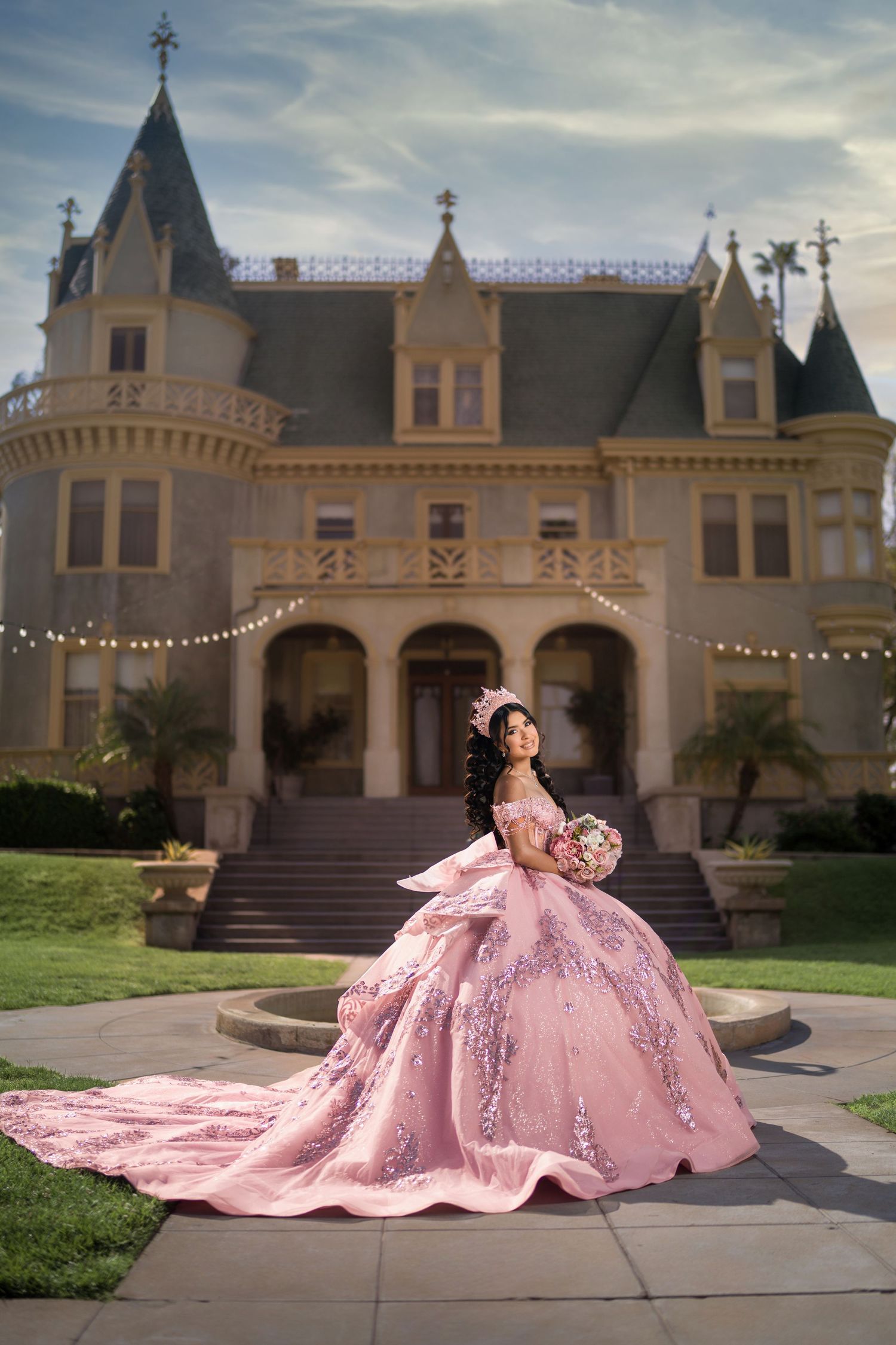 Young woman in pink quinceañera ball gown poses before a Victorian mansion with string lights.
