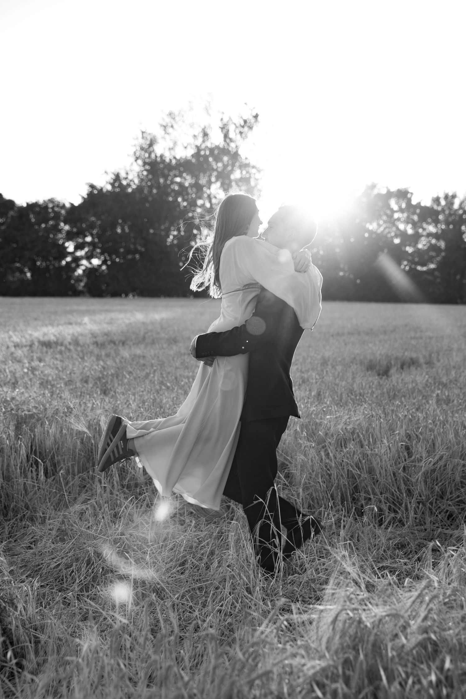 Black and white photo of groom lifting bride in sunlit field, romantic wedding portrait with lens flare.