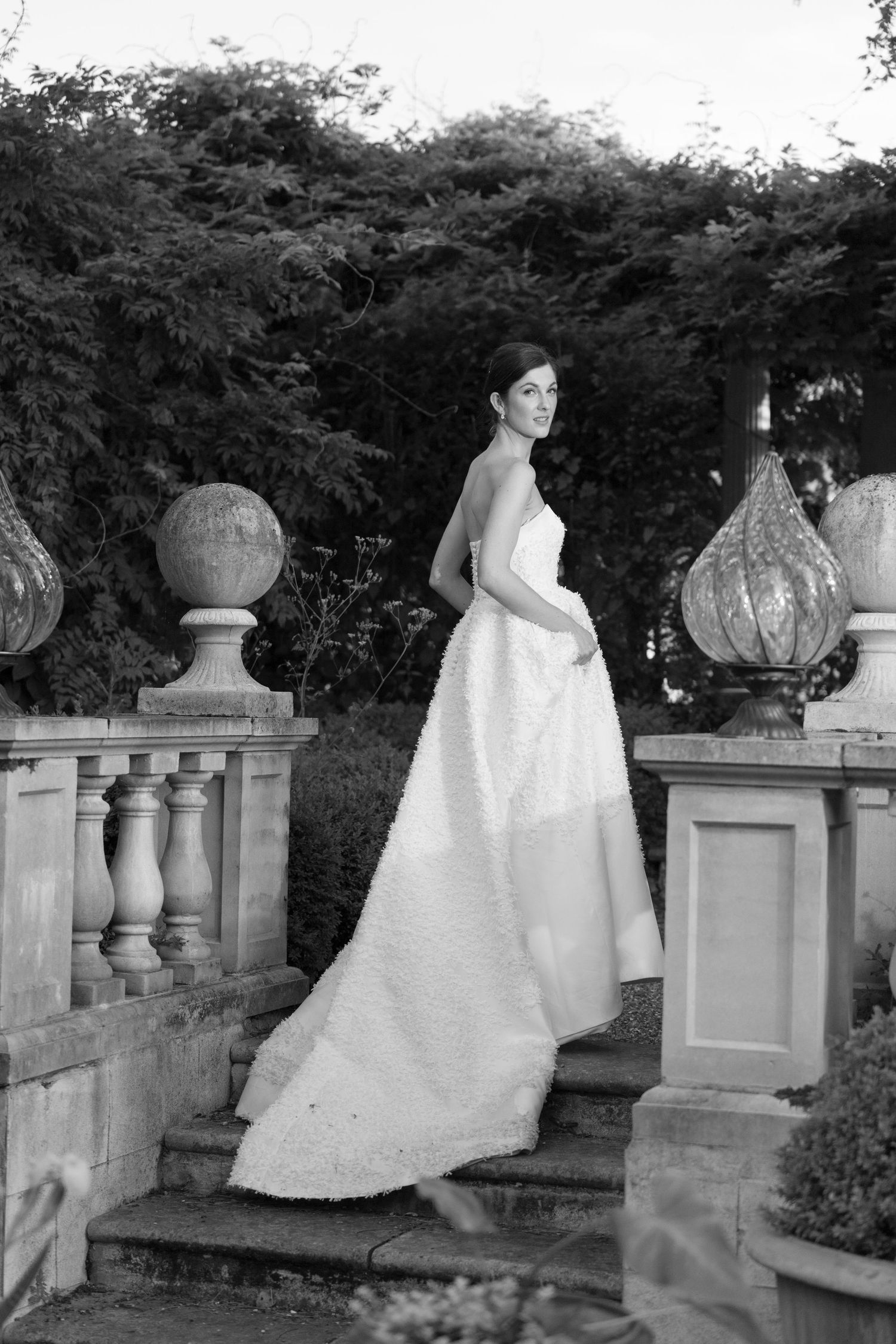 Bride in elegant white gown with long train poses on stone steps surrounded by lush gardens in black and white.