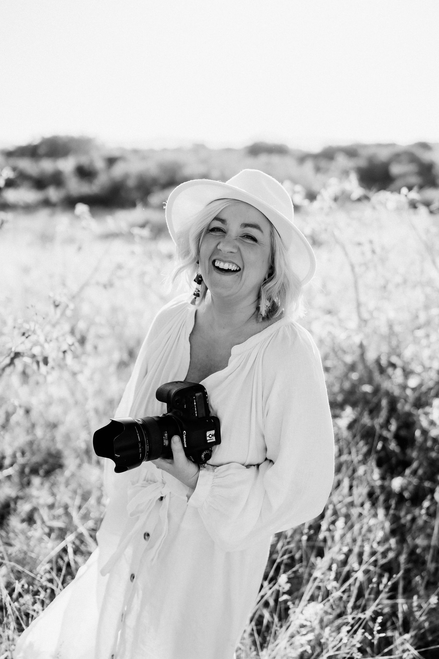 A photographer in a white dress and sun hat laughs joyfully while holding a camera in a sunny meadow.