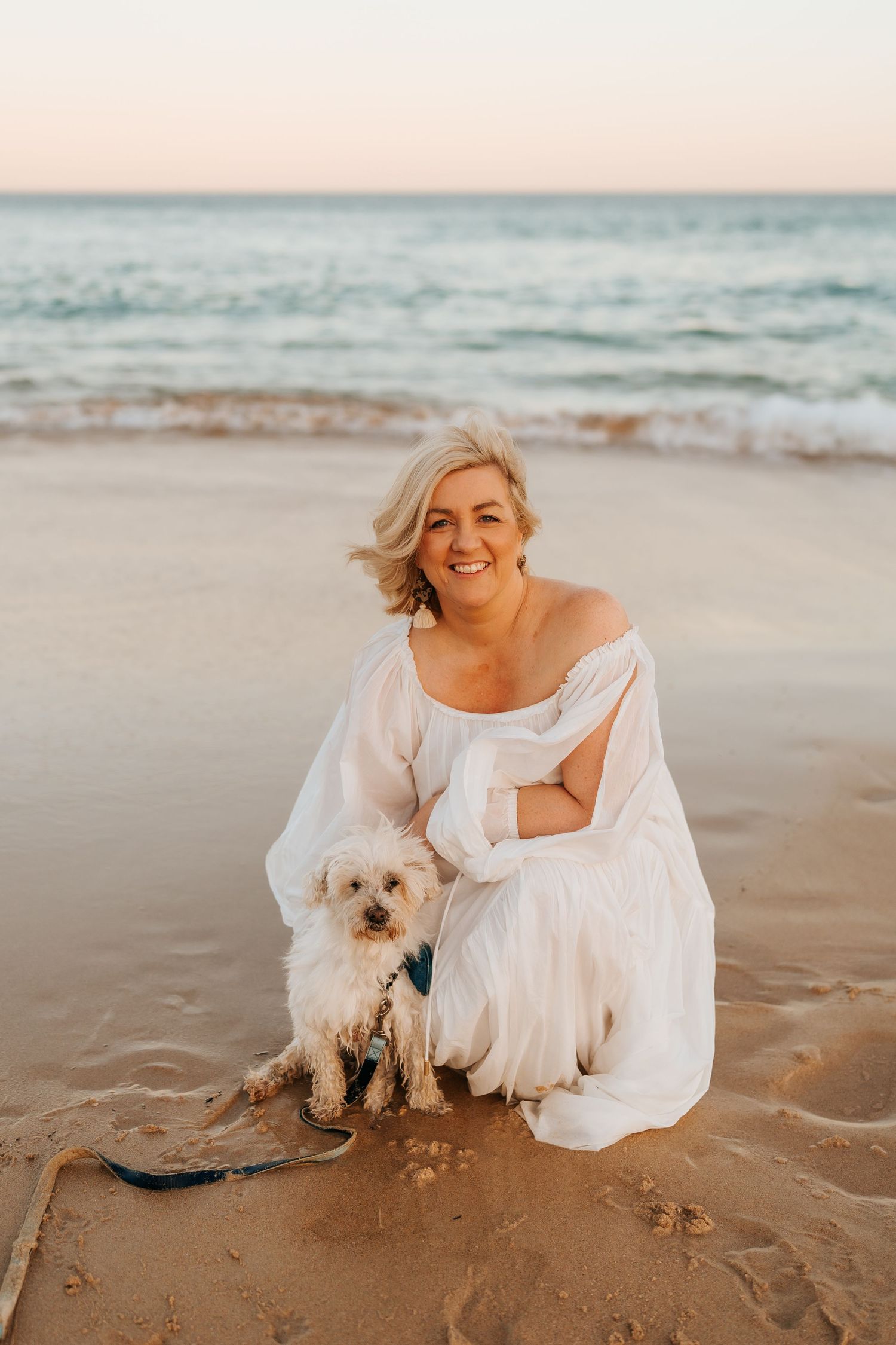A white-dressed figure poses with a small fluffy dog on a sunset beach.