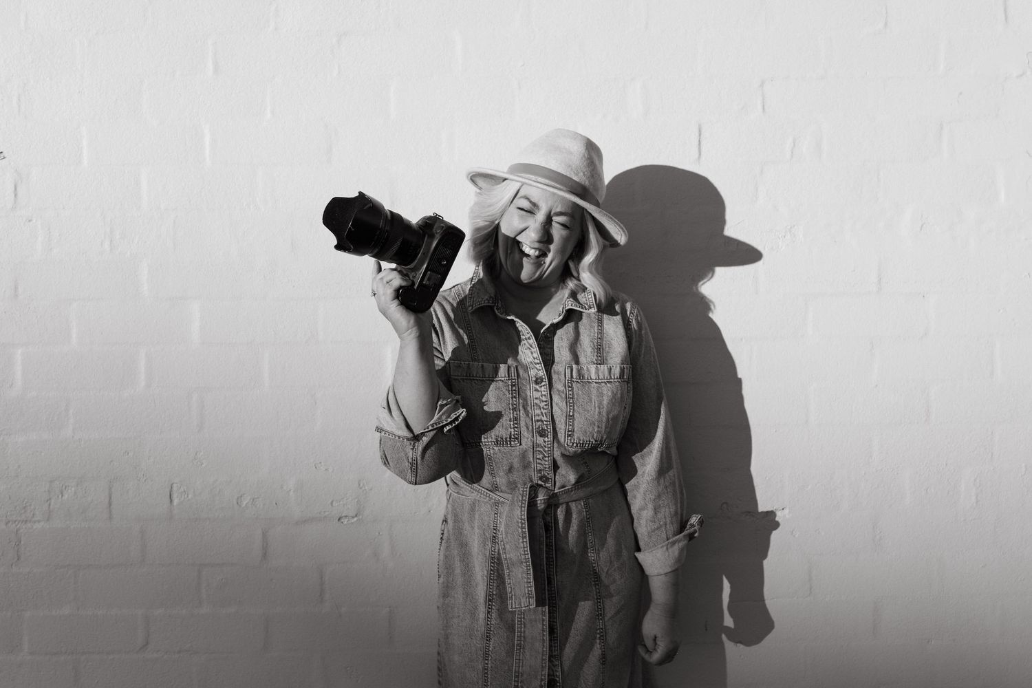 Happy woman in hat holding camera, laughing against white brick wall, black and white photography portrait.