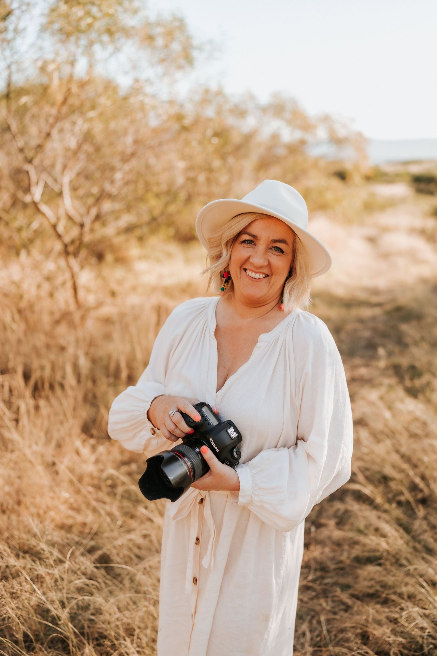 Photographer wearing white dress and hat smiles while holding professional camera in outdoor setting.