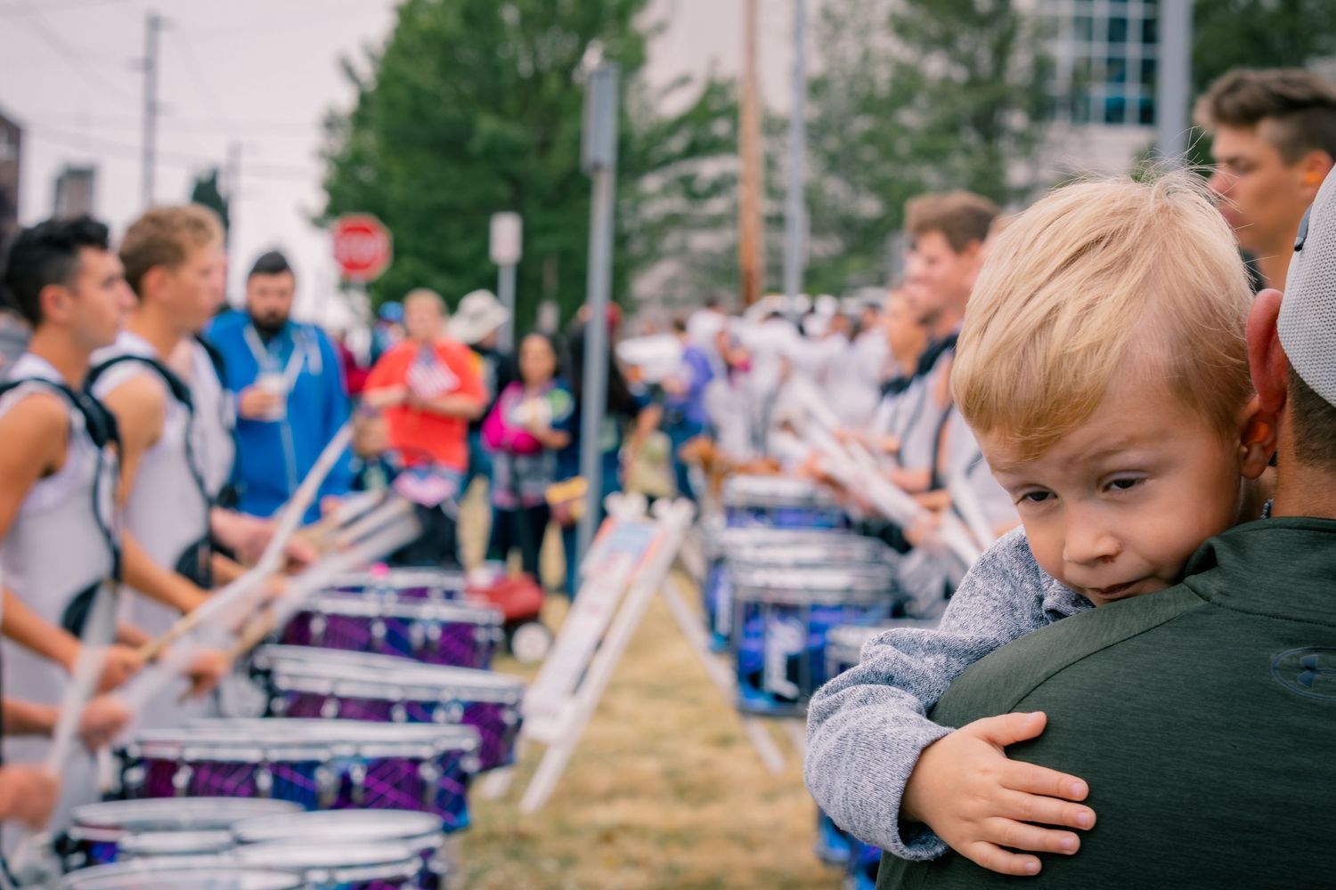 A young toddler rests on an adult's shoulder while a marching band drumline performs nearby outdoors.