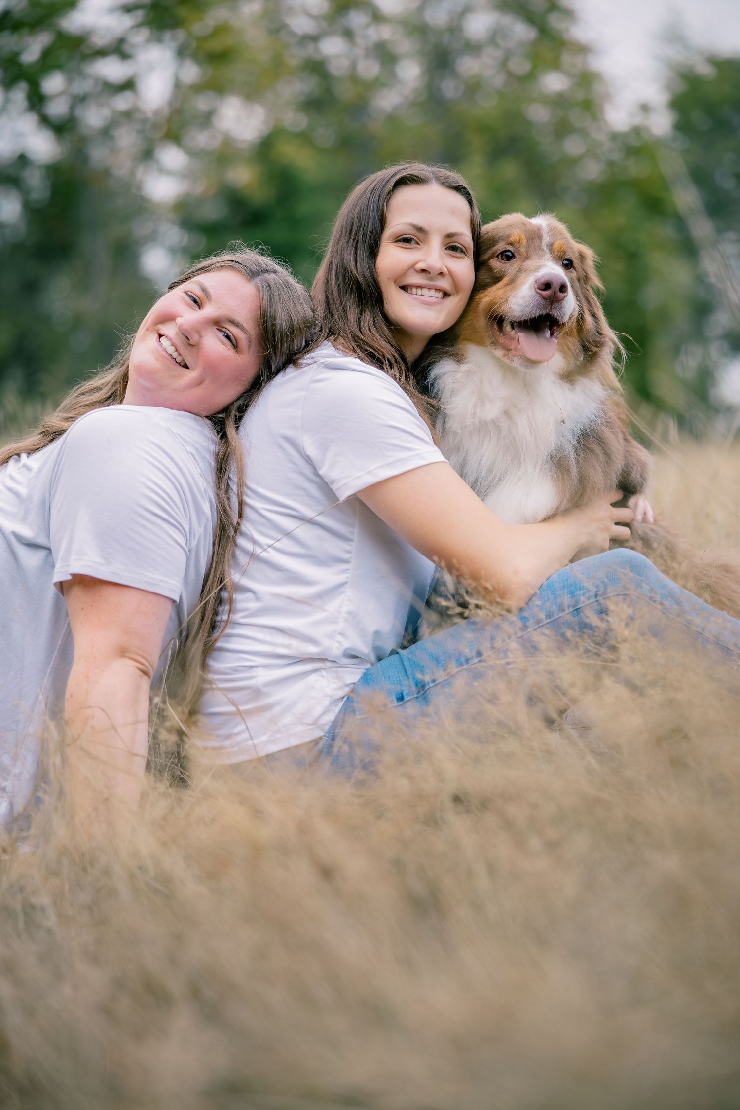 Two smiling women sit back to back in tall golden grass with an Australian Shepherd dog.