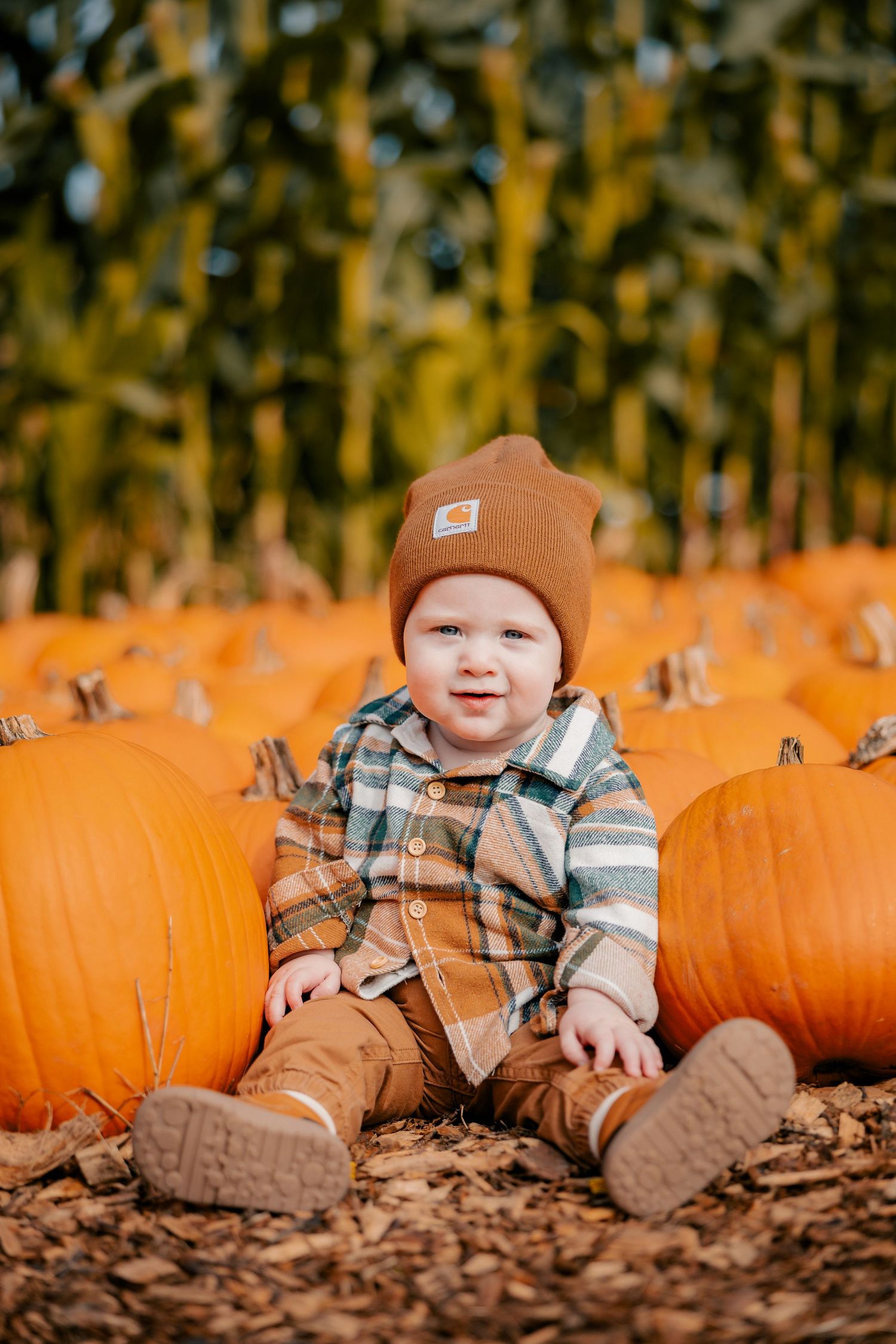 Smiling baby in Carhartt beanie and plaid overalls sitting among orange pumpkins at a fall pumpkin patch.