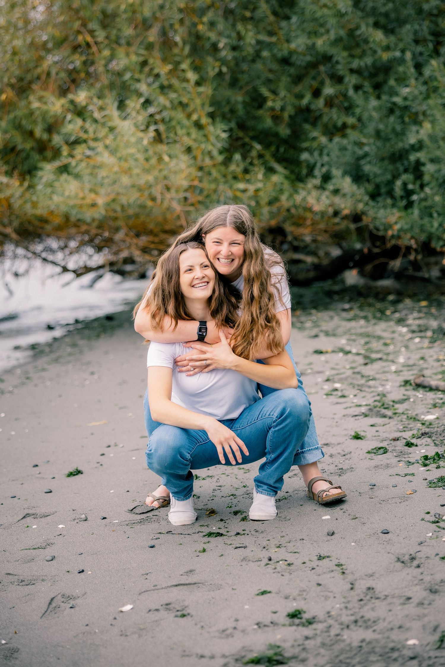 Two smiling young women embrace on a sandy beach surrounded by lush green foliage.