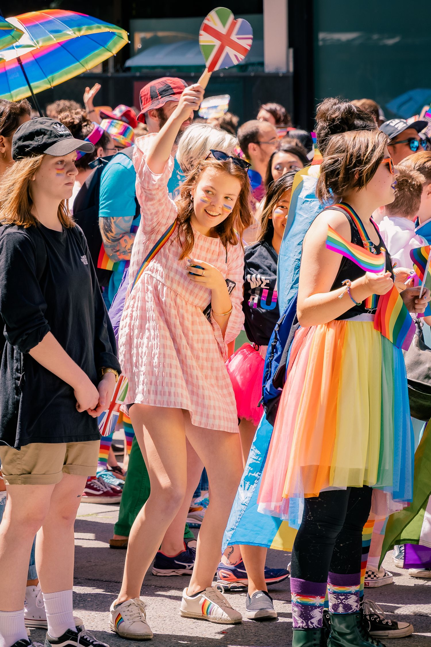 Joyful crowd at a Pride parade with colorful rainbow outfits, a heart-shaped flag, and rainbow umbrella on a sunny day.