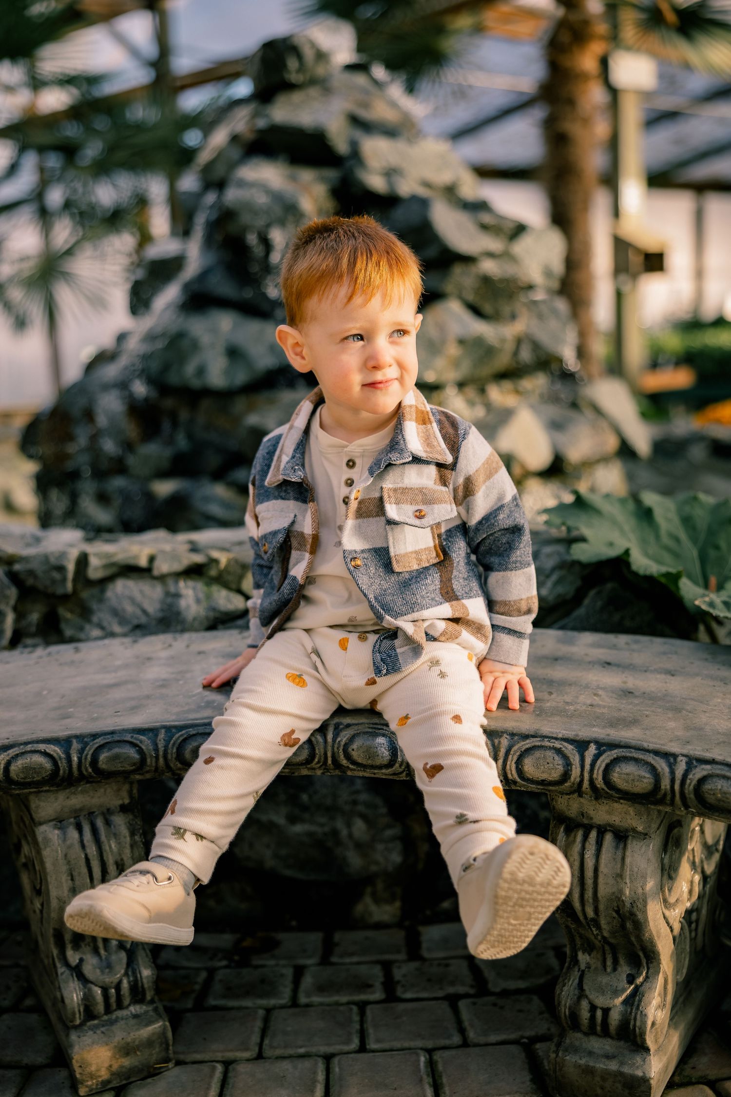 Cute red-haired toddler boy sitting on a stone bench wearing a striped jacket outdoors.