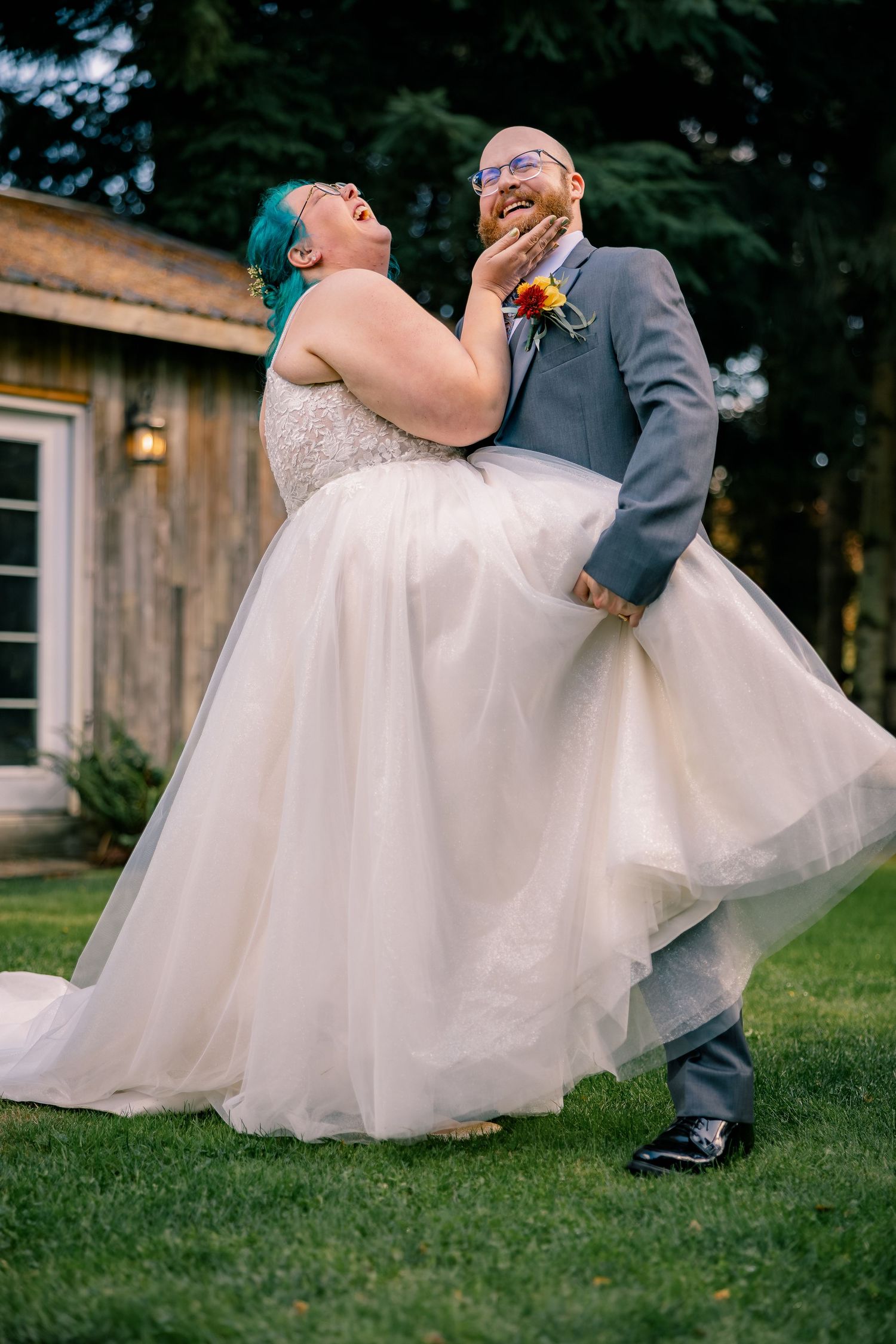 Laughing bride with blue hair playfully holds grooms face during outdoor wedding portrait on green lawn.
