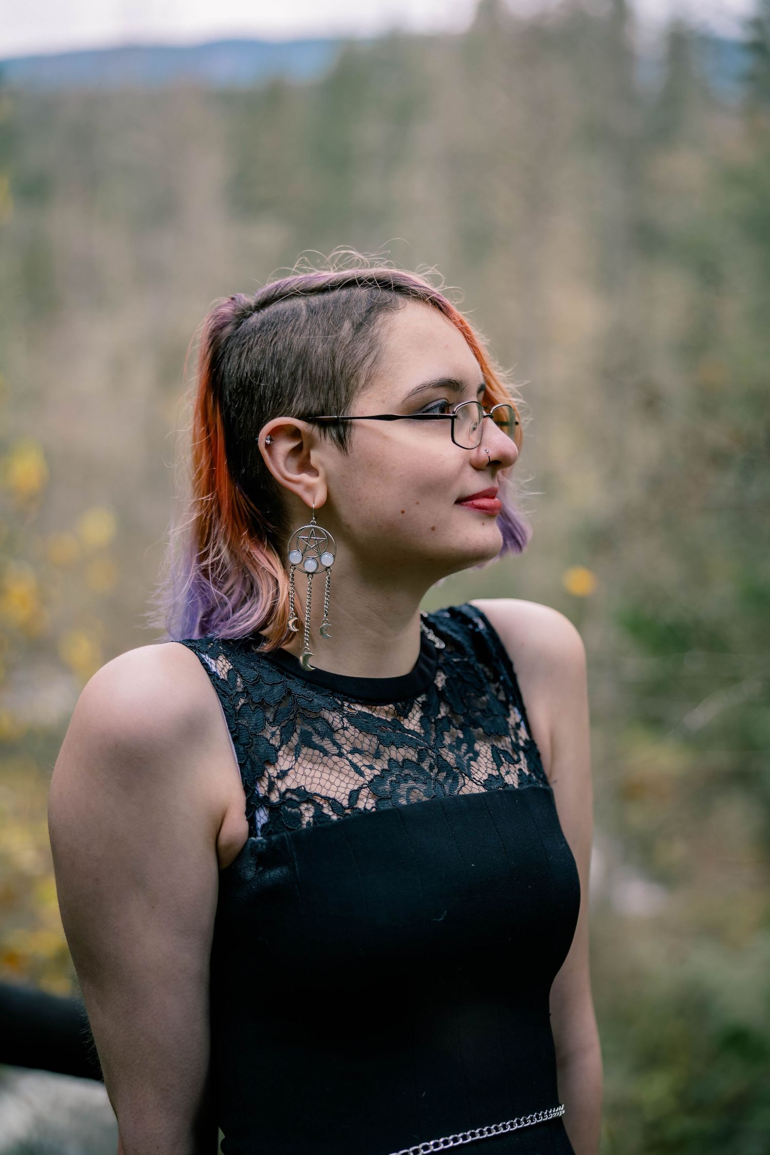 Side profile of a young woman with colorful hair, glasses, and dangling earrings in a black lace top outdoors.