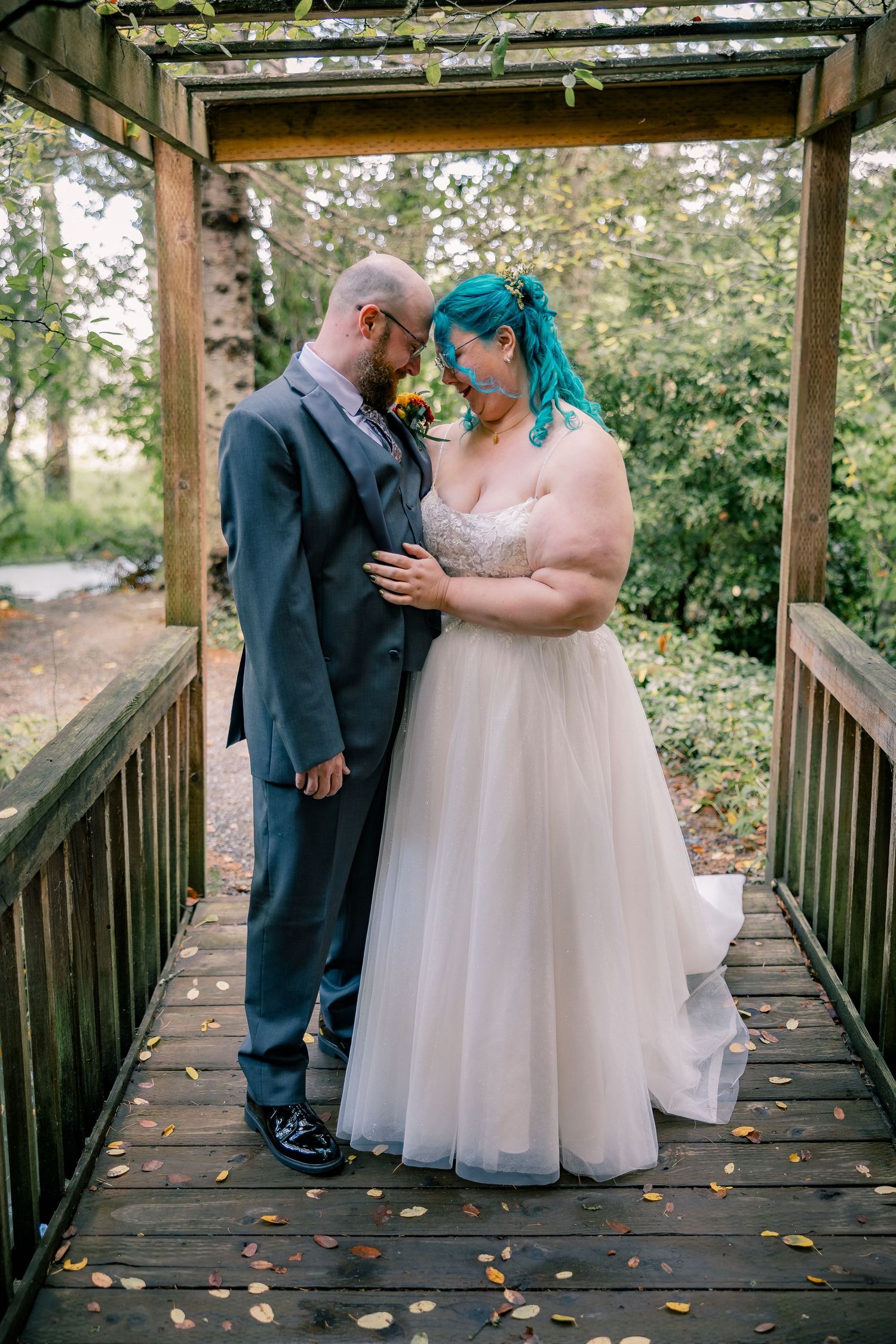 Bride with teal hair and groom share a tender moment on a wooden bridge in a garden.