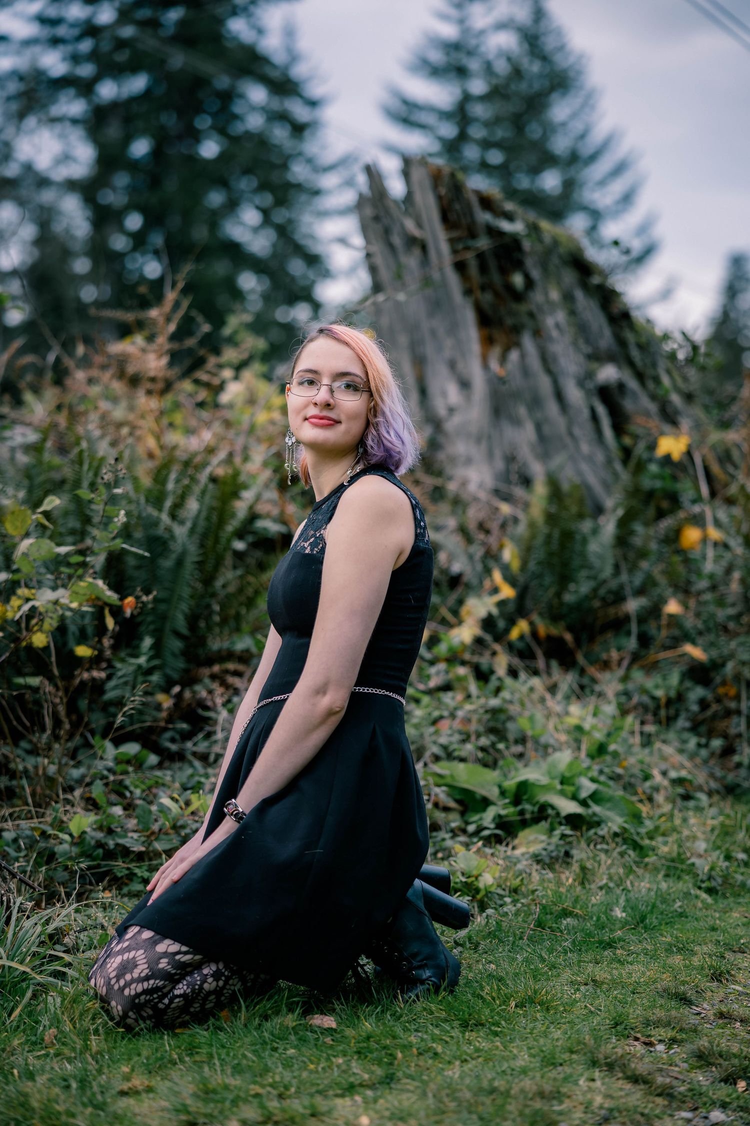 Young woman with purple hair in a black dress kneeling in a forest clearing near a tree stump.