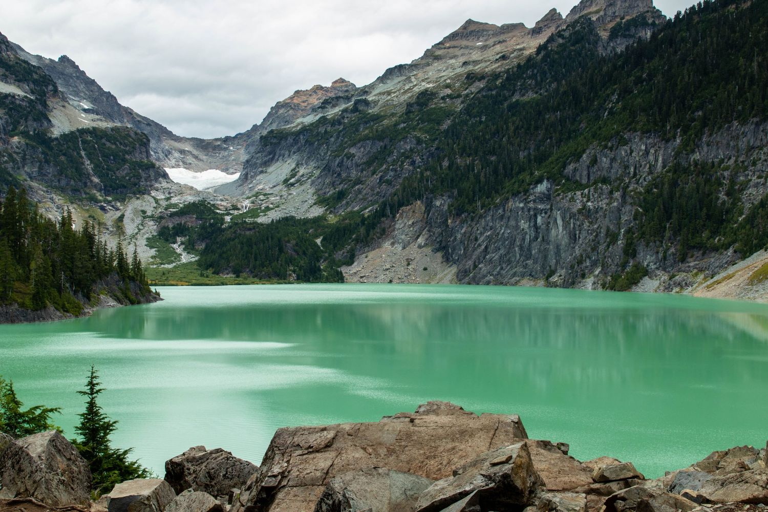 Stunning turquoise glacial lake surrounded by rocky mountains and evergreen trees under overcast skies.