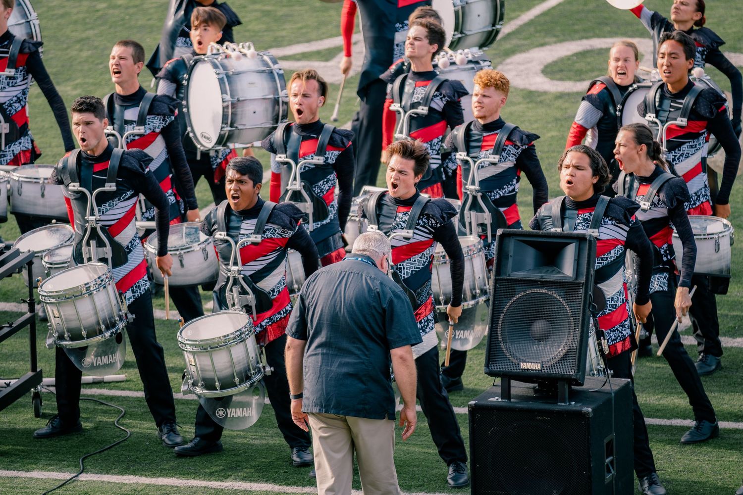 Marching band drummers perform in uniform on a football field while a conductor directs them.