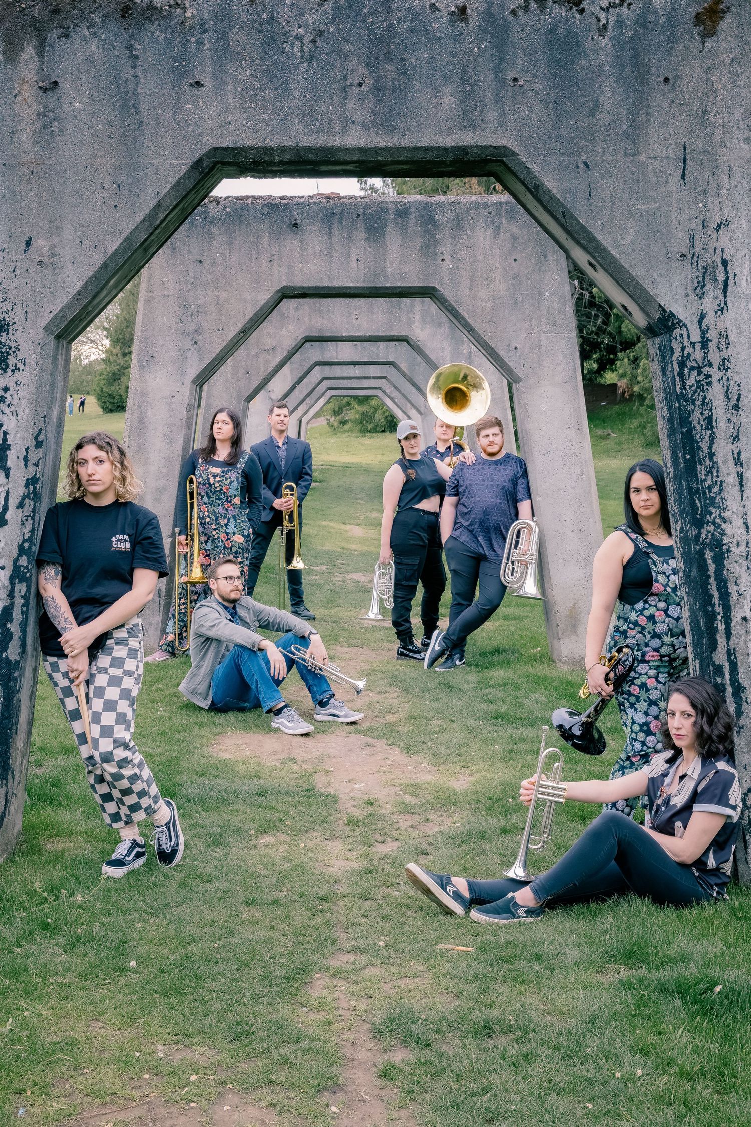 A brass band group poses artistically under concrete archway tunnels in a lush green outdoor park.