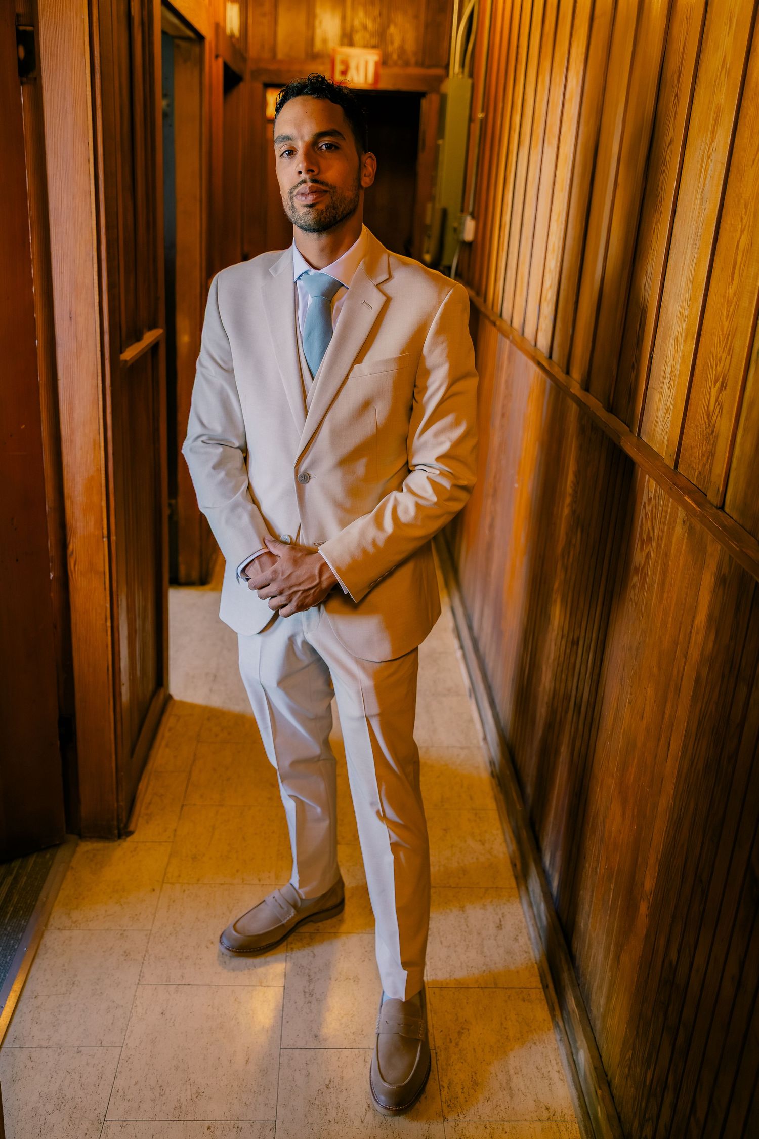 Stylish man in a beige suit and blue tie poses confidently in a warmly lit wood-paneled hallway.