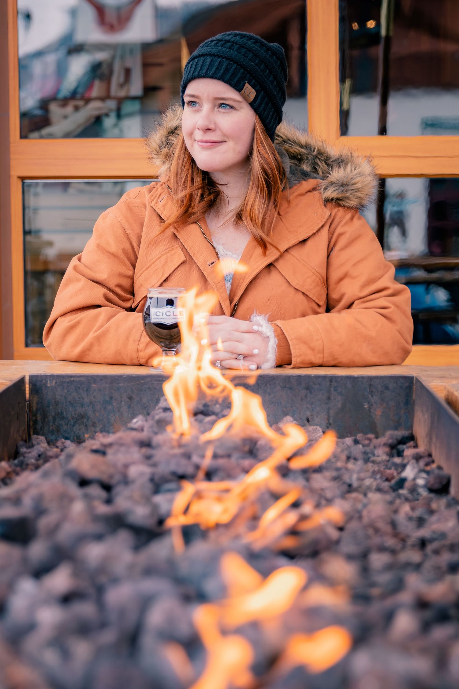 A woman in an orange jacket and black beanie holds a dark beer glass beside an outdoor fire pit.