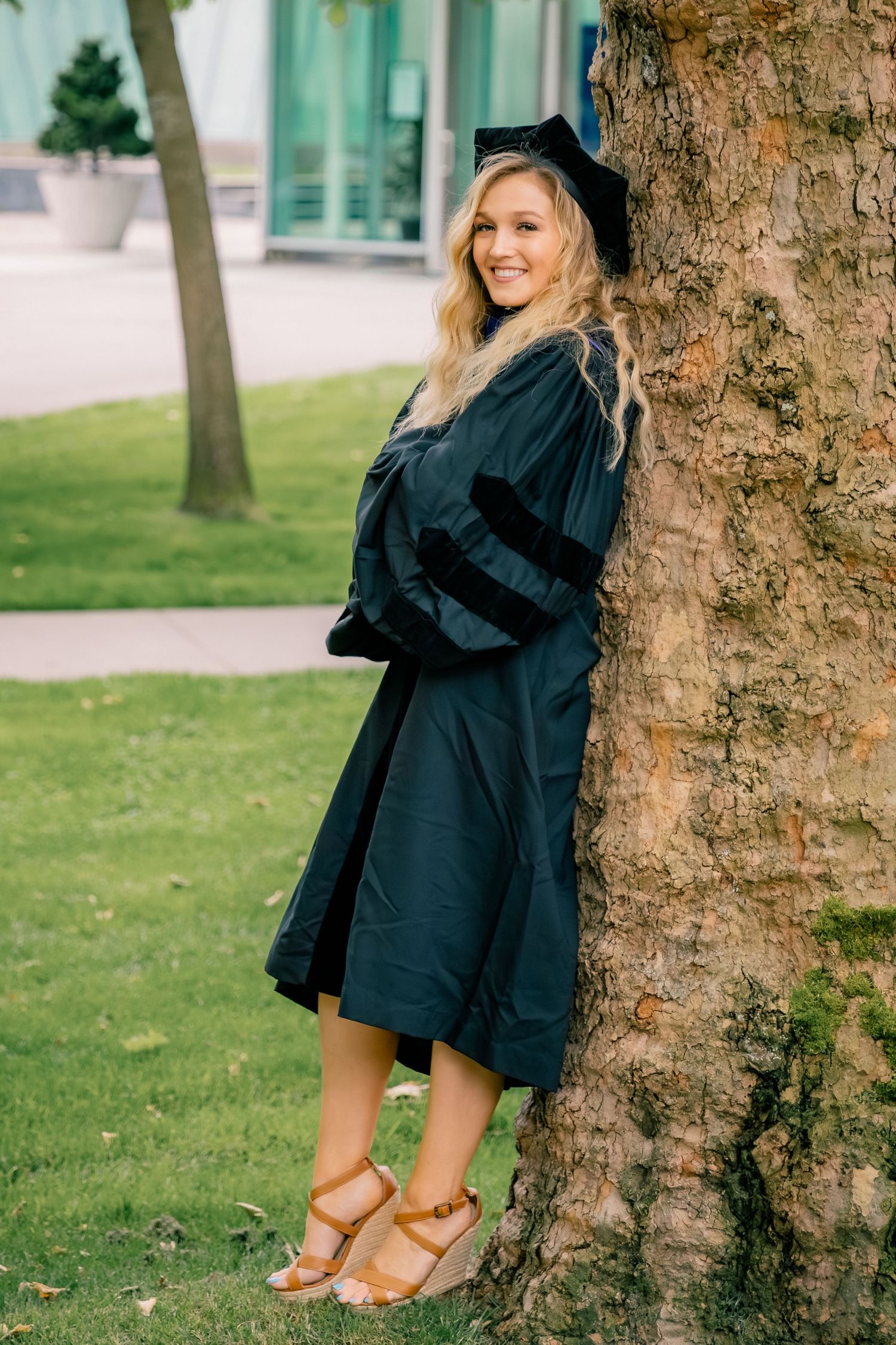 A smiling blonde woman in a black doctoral graduation gown and cap leans against a tree outdoors.