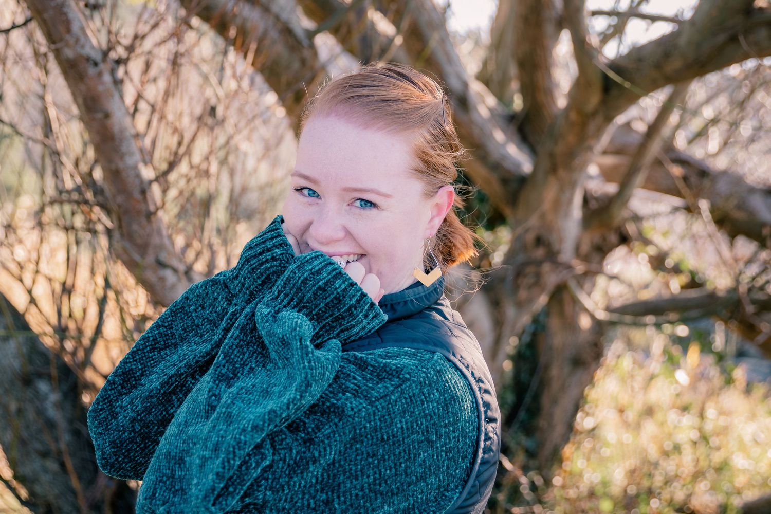 A young redheaded woman smiles warmly while wrapped in a teal sweater near bare winter tree branches.