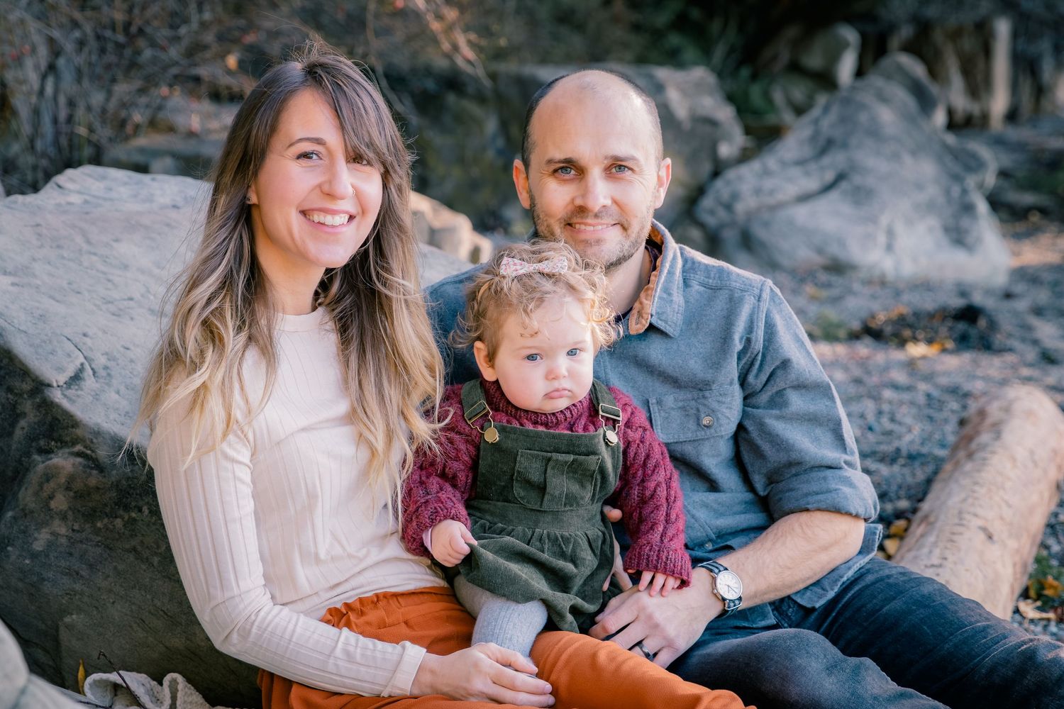 Smiling young family with baby girl in green overalls posing on rocks in nature.