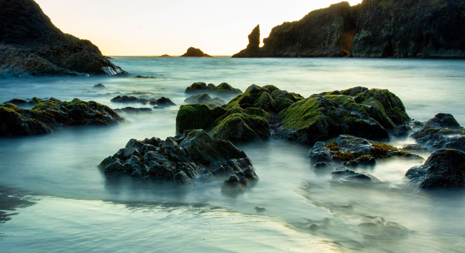 Mossy rocks on a misty coastal beach at sunset with calm ocean waves and a sea stack silhouette.