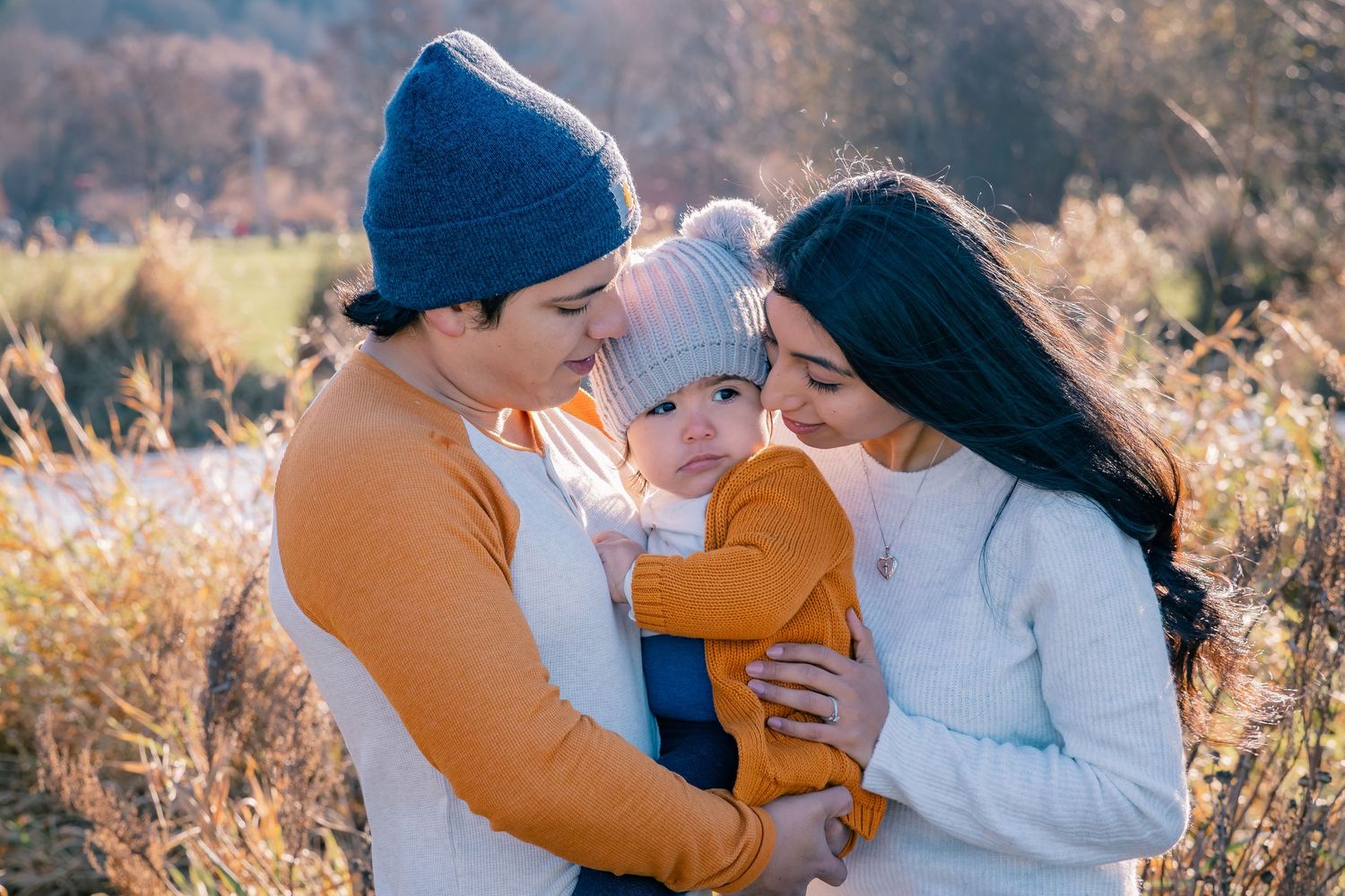 Young couple in autumn field tenderly holding their baby wearing matching warm hats outdoors.