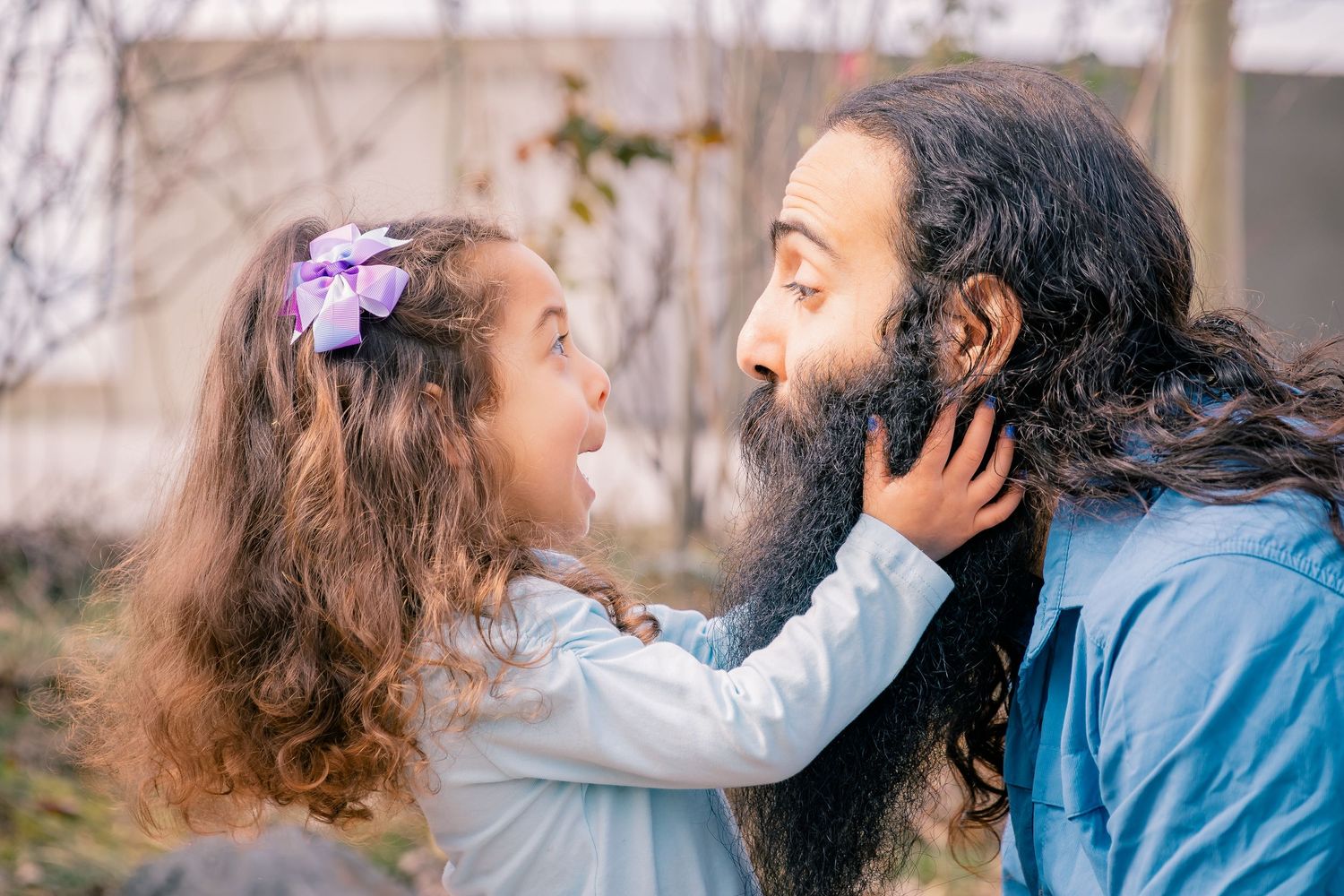 Adorable curly-haired girl laughing while touching her father's long beard in a garden.