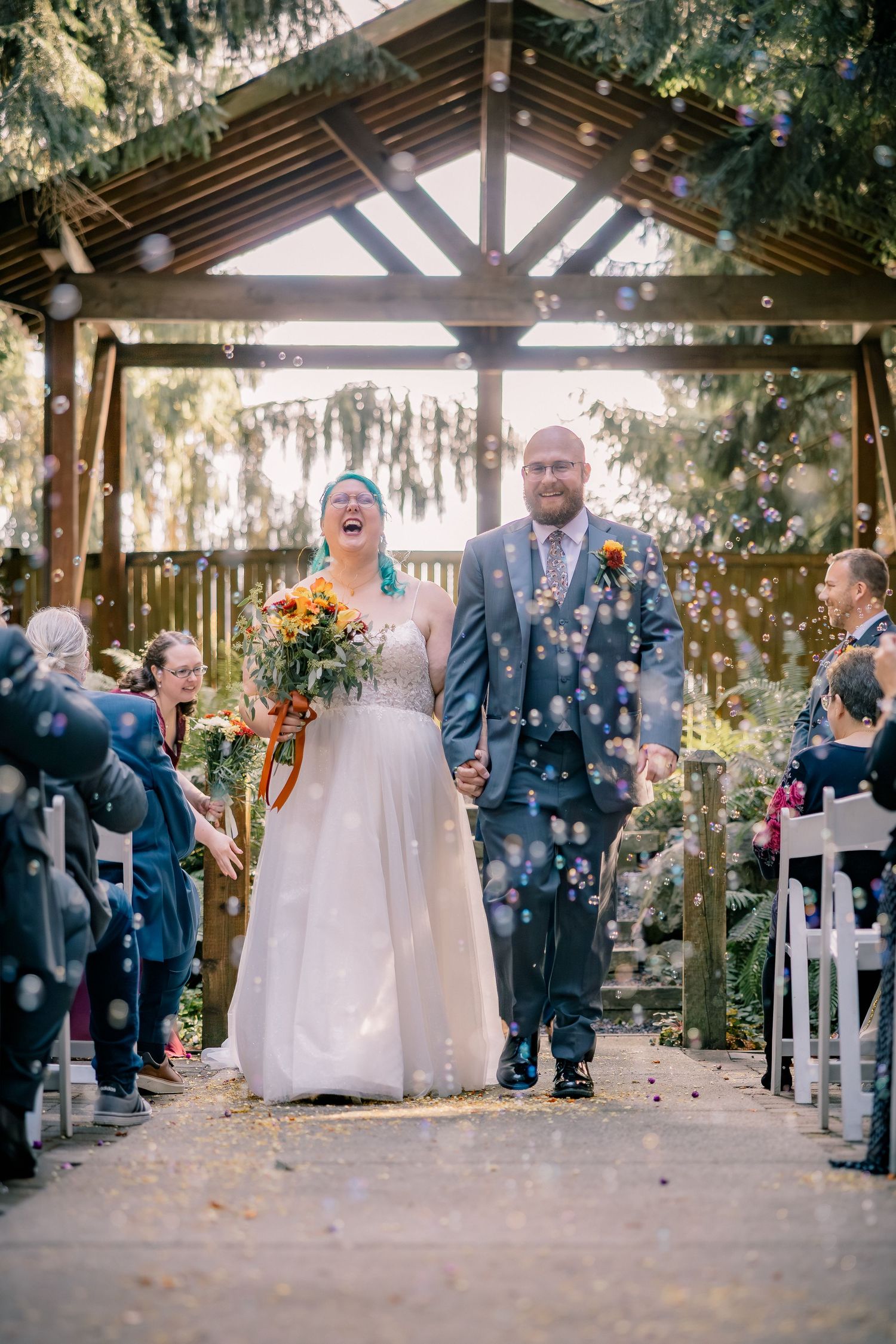 Joyful newlyweds walk down the aisle surrounded by bubbles at an outdoor garden wedding ceremony.