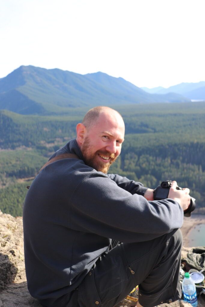 Smiling bearded man sitting on a mountain overlook holding a camera with scenic forest and peaks behind him.