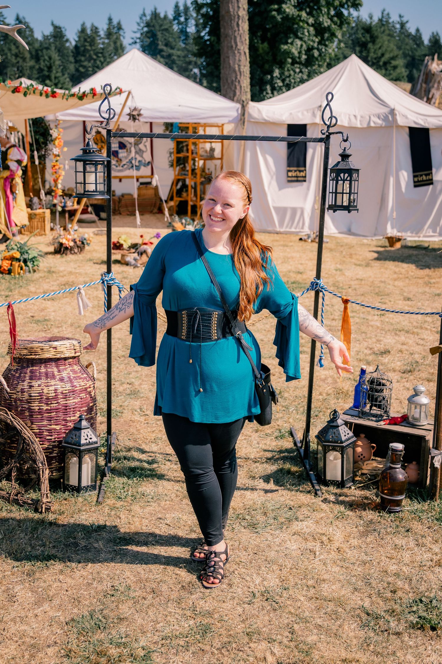 A smiling redhead woman in a teal belted tunic walks through a medieval fair marketplace on a sunny day.