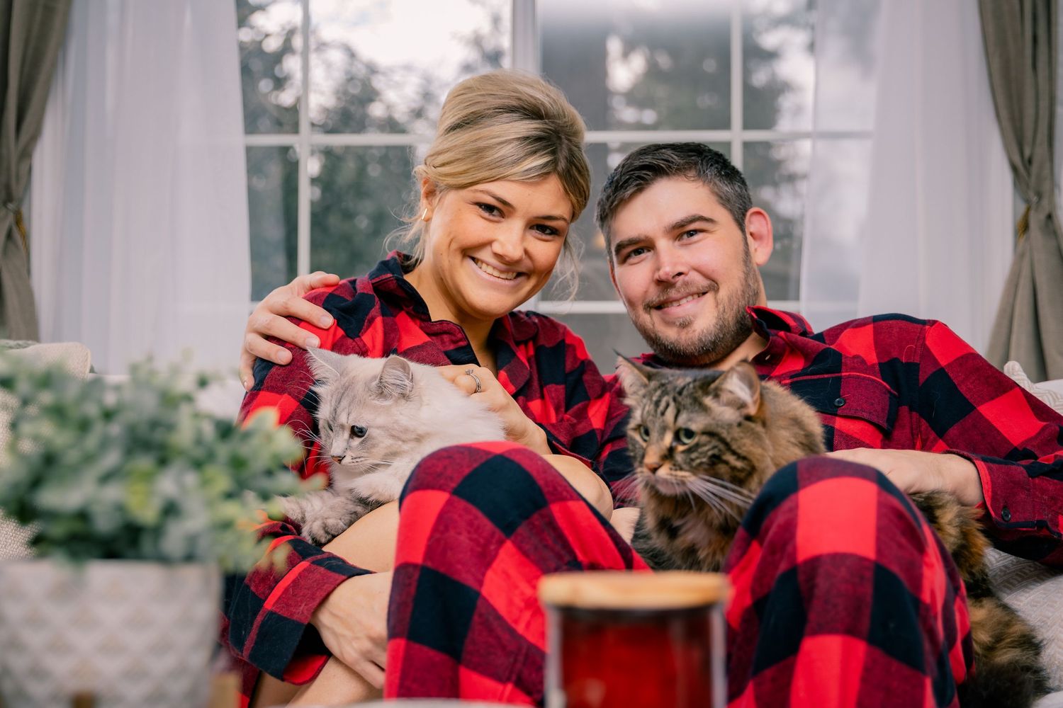 A smiling couple in matching red plaid pajamas cuddles together on a couch with two fluffy cats.
