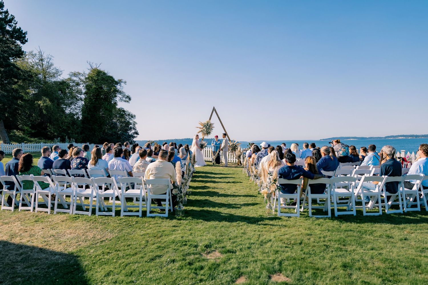Elegant outdoor waterfront wedding ceremony with guests seated on white chairs facing a triangular arch.