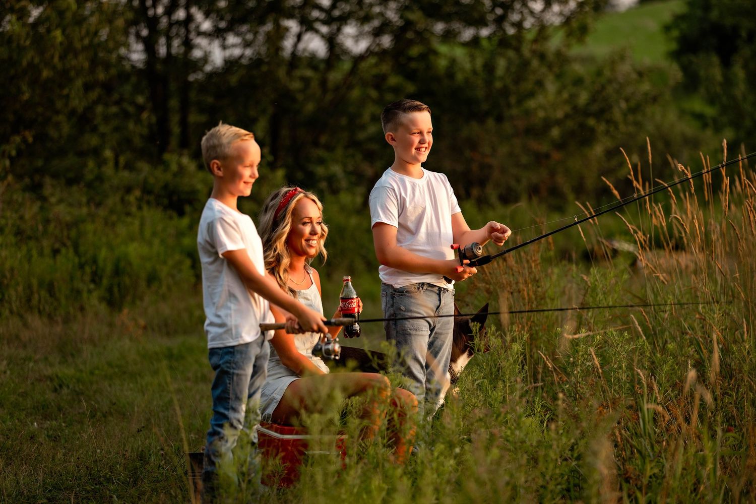 Mom sitting on an old cooler drinking coke while her kids fish at the pond
