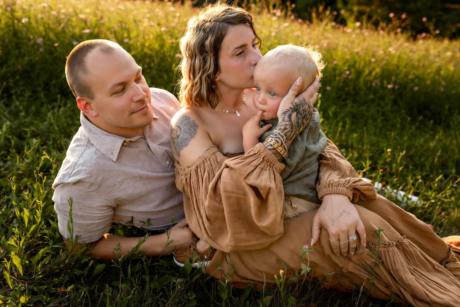 Parents sitting in the grass with their toddler while mom kisses his head in Central NY
