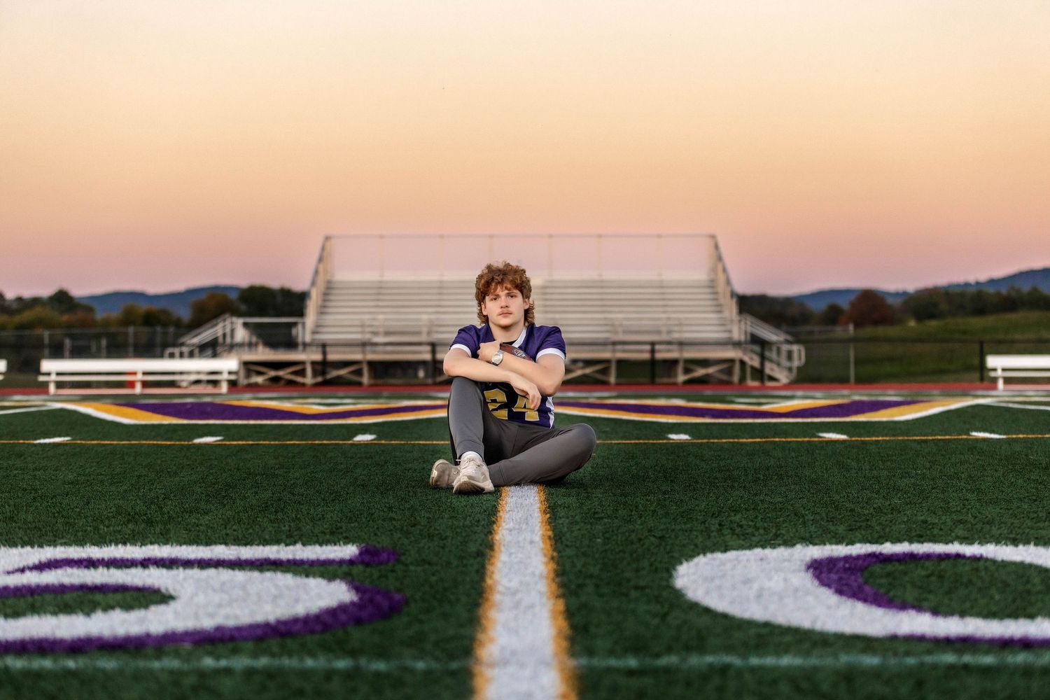 Senior boy sitting on 50 yard line in football jersey holding football with a sunset background in Central NY