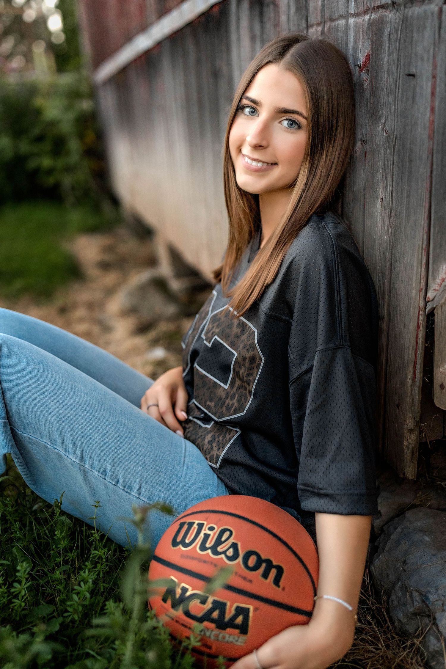Senior girl with basketball and jersey leaning against rustic wood building in Central NY