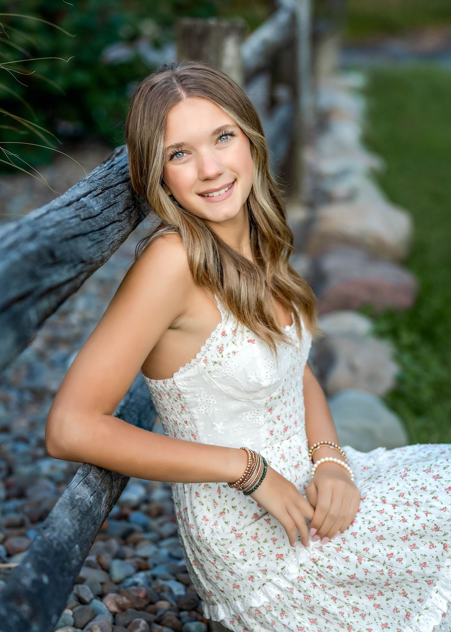 senior girl sitting on rocks wearing a dress and leaning against a wooden fence