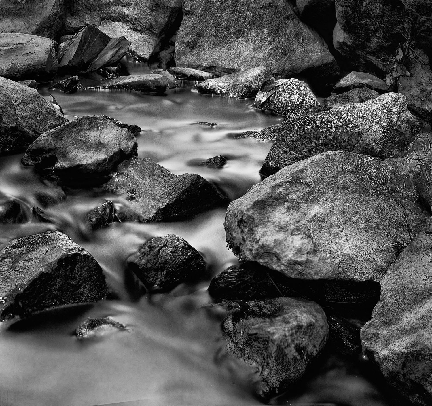 Long exposure black and white photograph of flowing water cascading over smooth rocks in a stream.
