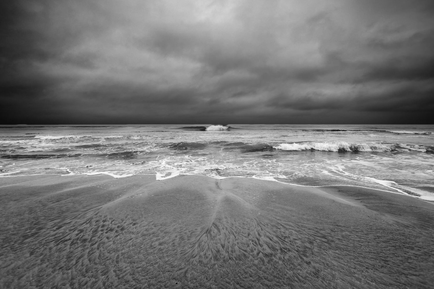 Dramatic black and white seascape shows rolling waves under stormy skies with dramatic cloud formations.
