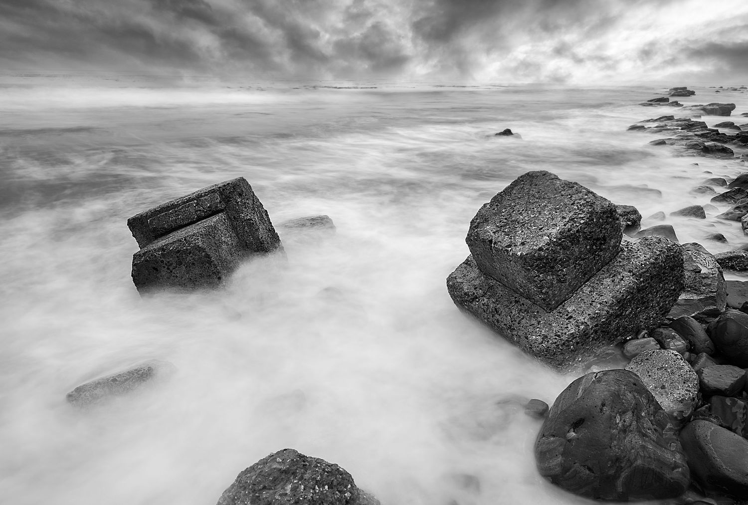 Long exposure seascape photograph captures waves flowing around dark rocks under dramatic cloudy sky.