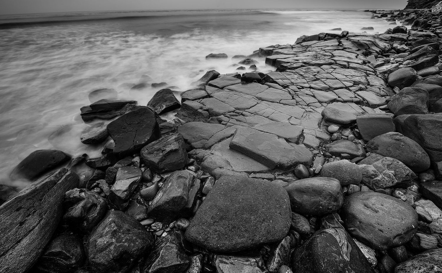 Dramatic black and white landscape of rocky shoreline with crashing waves and moody sky at sunset.