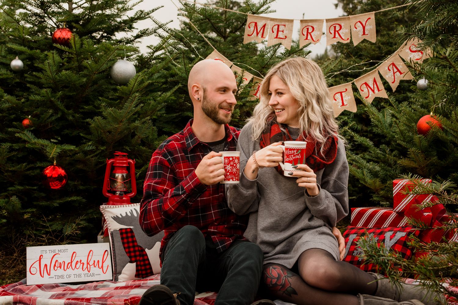 A couple hold mugs of hot chocolate and smile at each other as they sit in a Christmas decorated wonderland.
